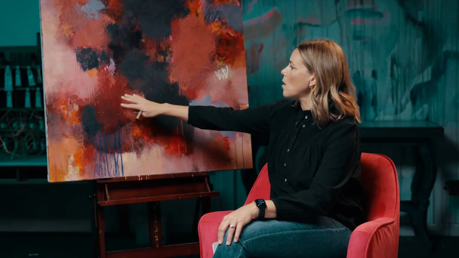 Casey Wakefield pointing to layered red, pink and charcoal areas of her large abstract painting on an easel