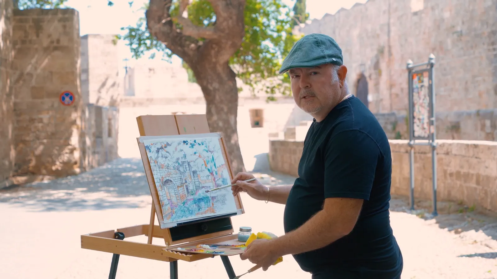 John Milan painting at a portable easel outside a medieval stone wall under large trees