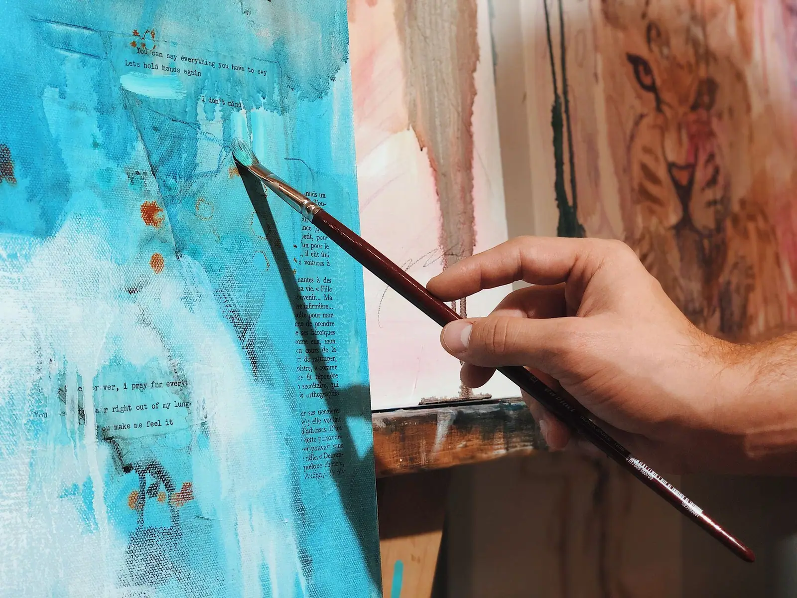 Hand holding a brush over a blue mixed media painting with translucent washes