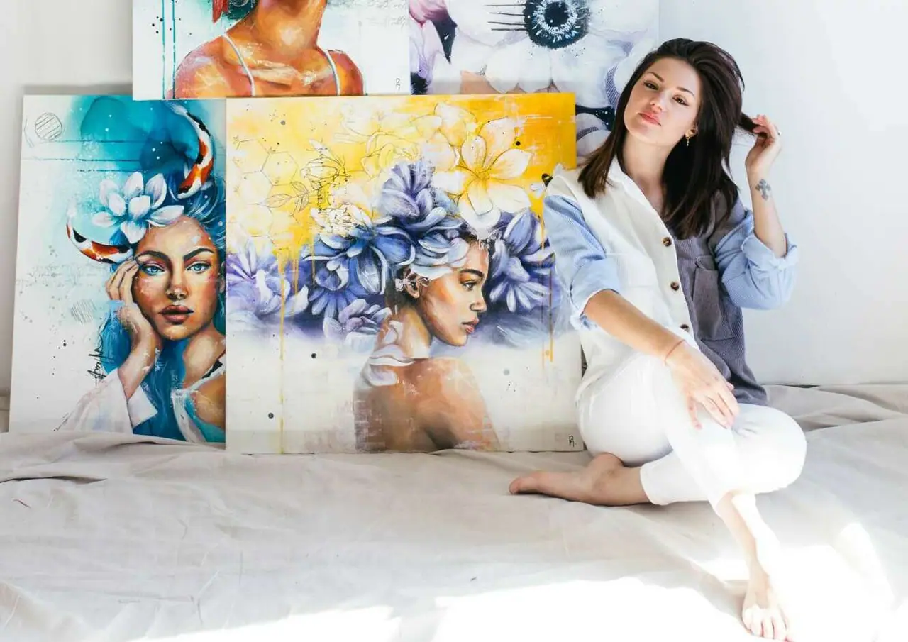 Artist seated in a gallery beside several large colorful portrait paintings
