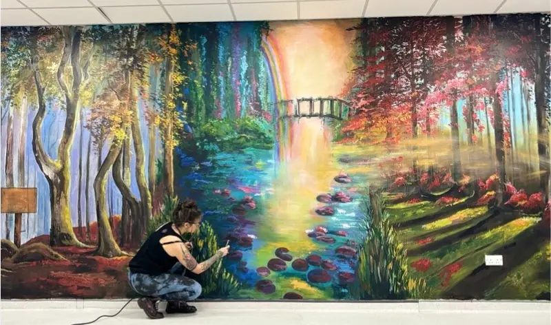 Lisa Cunningham working on a mural of a forest stream with a bridge and trees with pink and red leaves