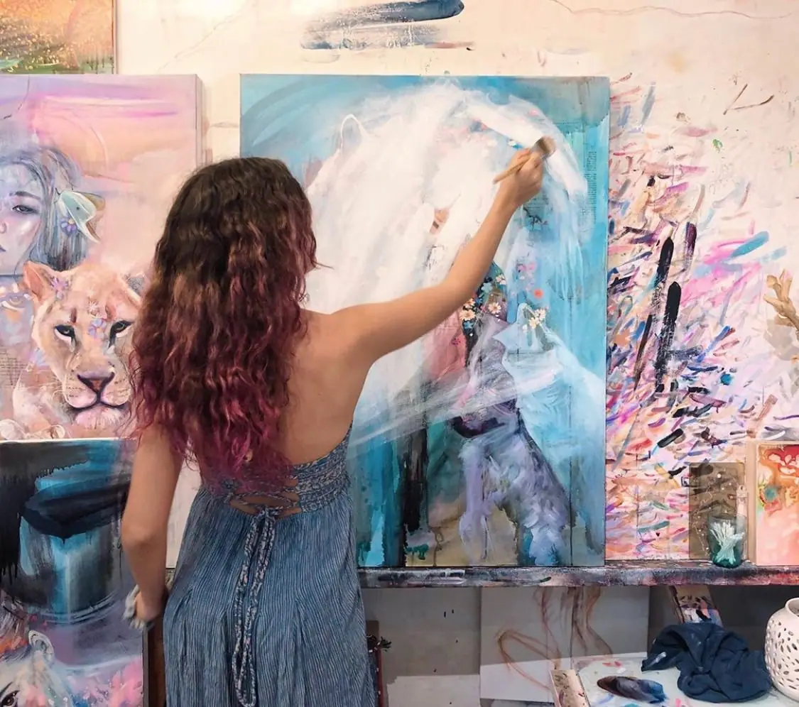 Artist brushing pale pink, blue, and cream oil paint over an old oil painting to create a fresh surface