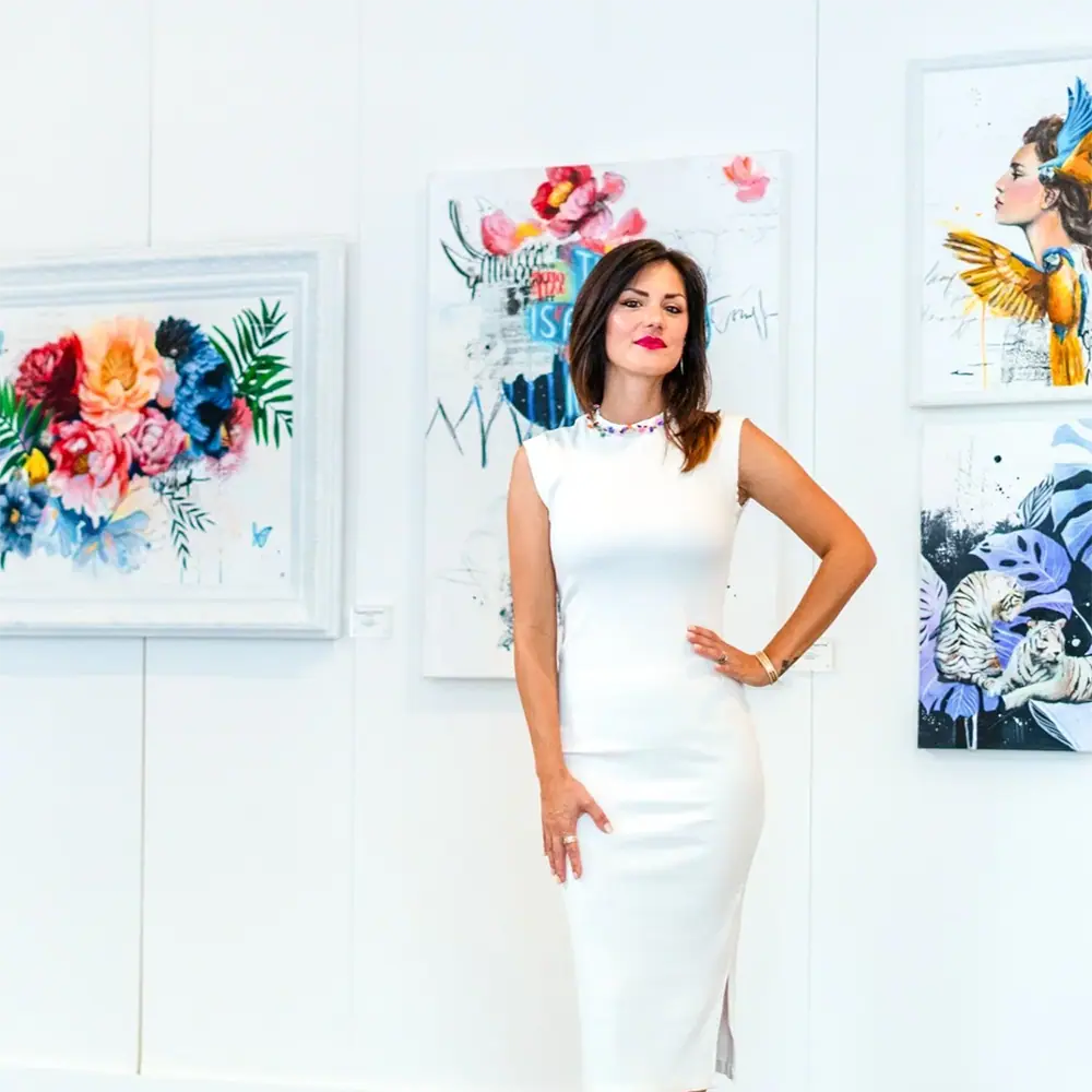 Artist in a white dress standing in a gallery with colorful artwork on the walls