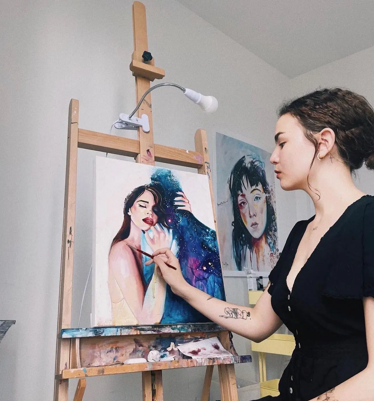 An artist painting a portrait on an easel lit by a clamp lightbulb fixture beside the canvas