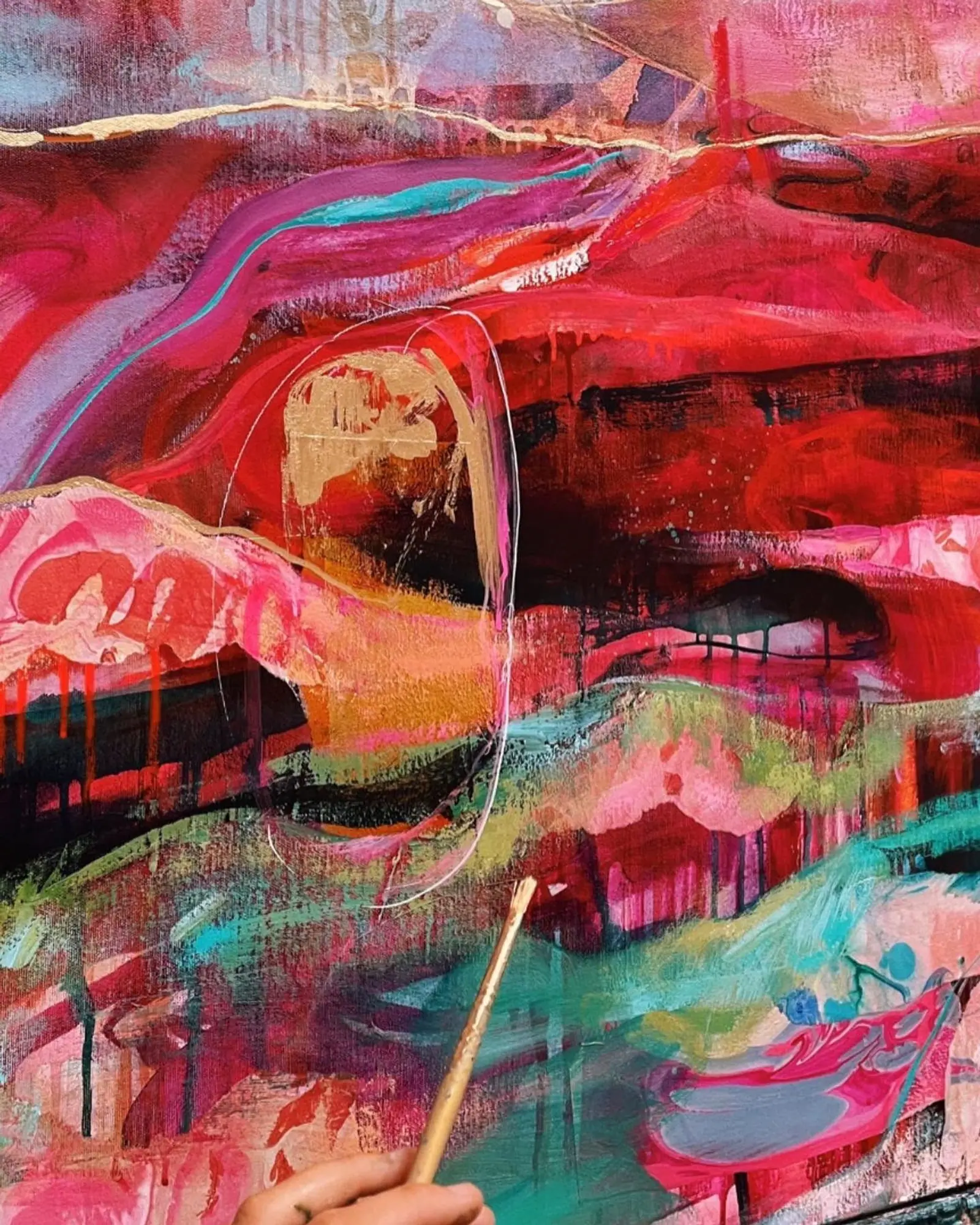Hand holding a paint brush in front of a colorful abstract canvas in red, blue, and gold