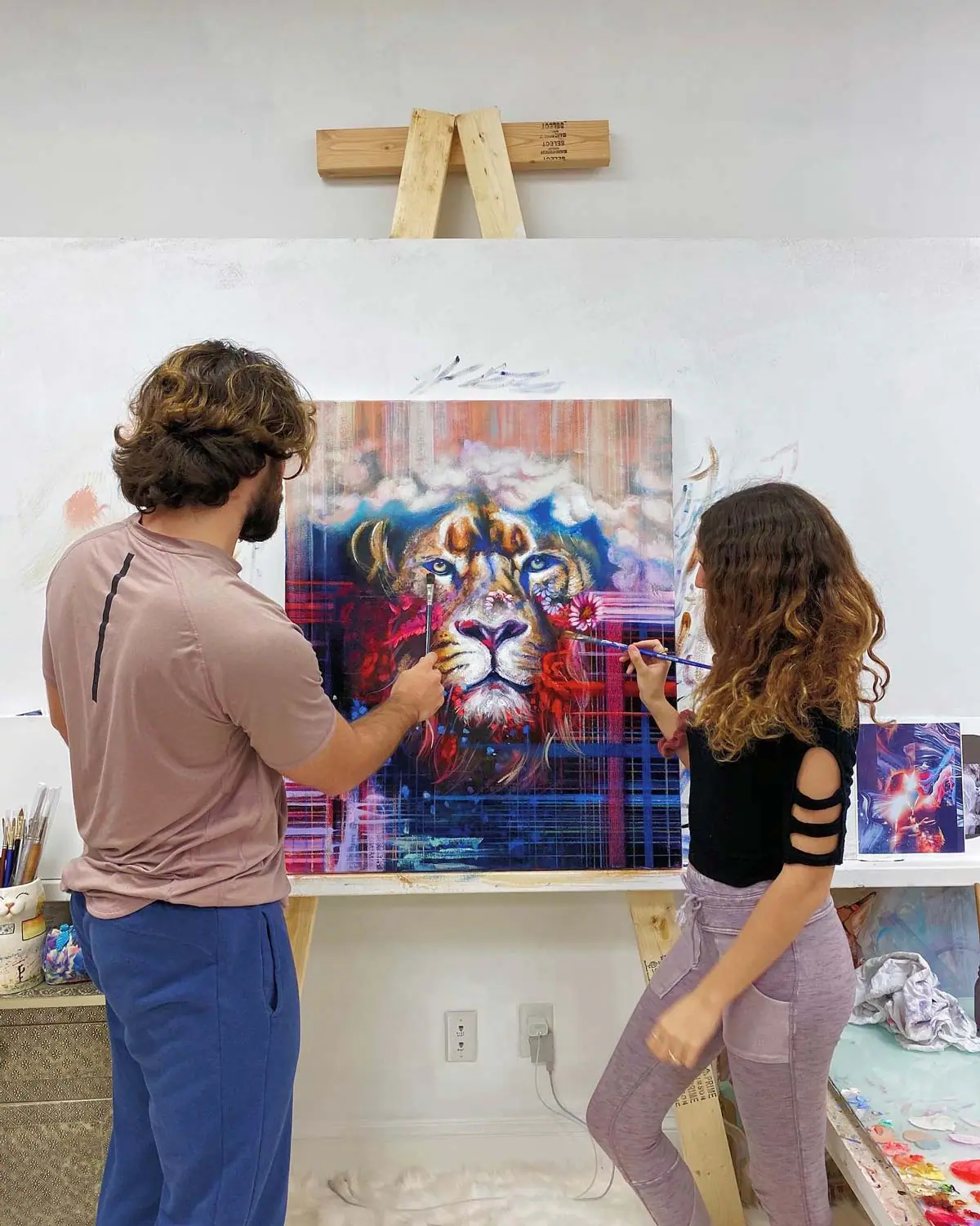 Two artists painting a large colorful lion together on an easel in a studio