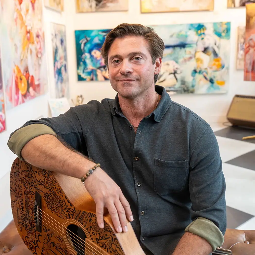 Gallery director Jock Armour seated in a gallery holding a guitar