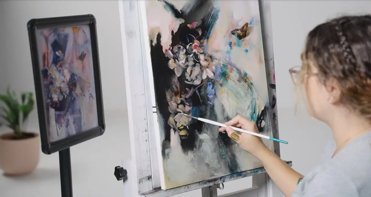 Artist painting an abstract flower on canvas with a reference image on a stand beside her