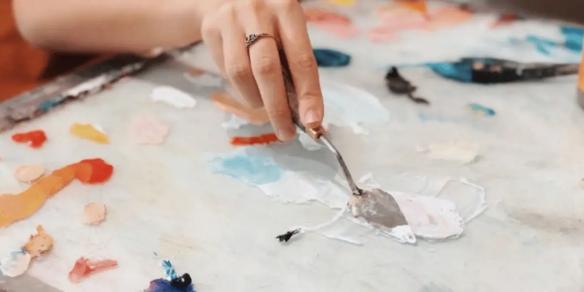 Hand holding a palette knife over a palette loaded with many colors of paint