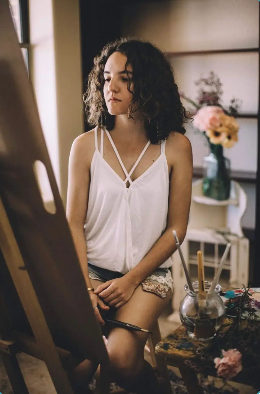 Artist Dimitra Milan seated at her easel working on a painting in the studio