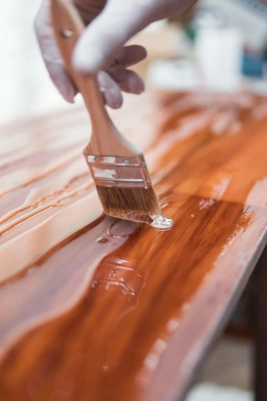 A soft brush spreading clear varnish across a wood surface in long even strokes