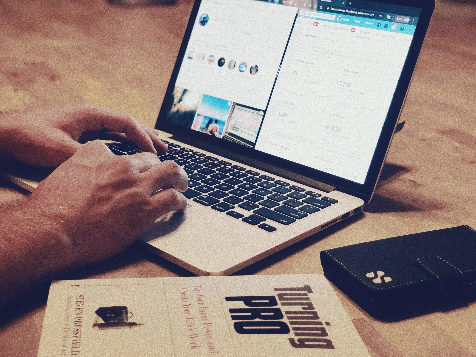 Artist working on a laptop at a wooden table with a book and phone, building an online portfolio
