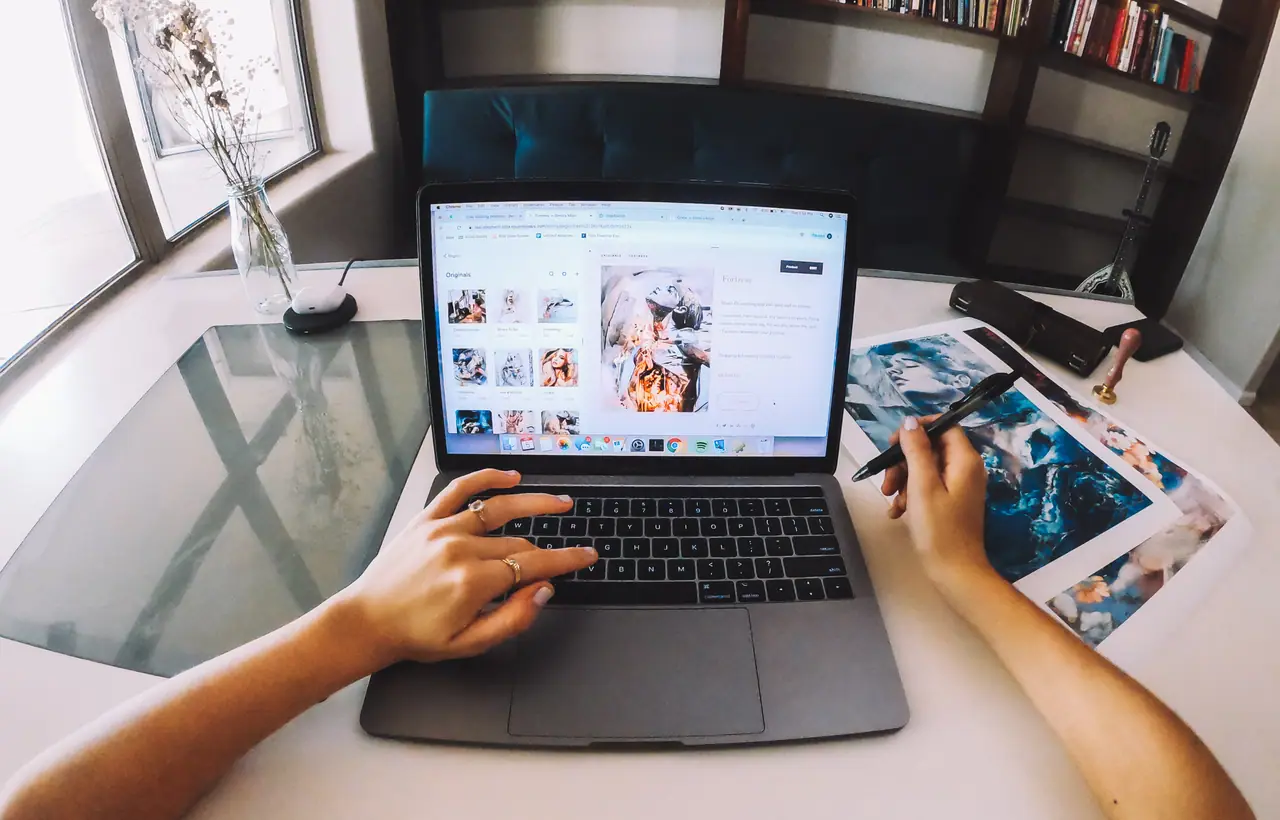 A hand working on a laptop next to a blue abstract painting on the table