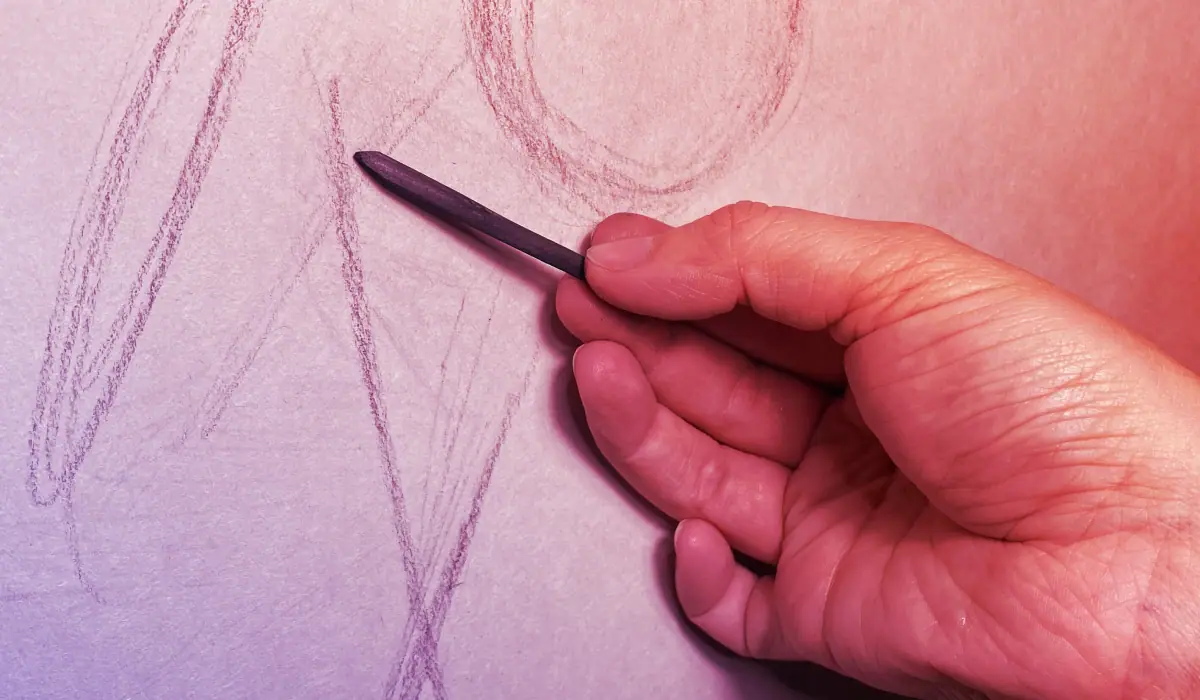 A hand holding charcoal loosely in front of a sheet of drawing paper