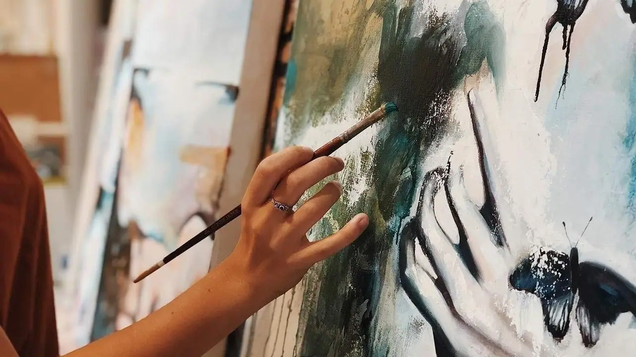 A hand holding a brush to a large earth toned canvas painted with a hand and a butterfly