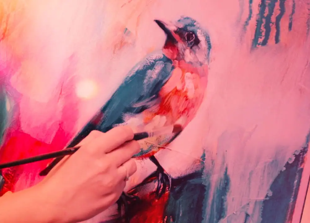 Artist's hand layering pink and blue paint over a bird in a mixed media painting