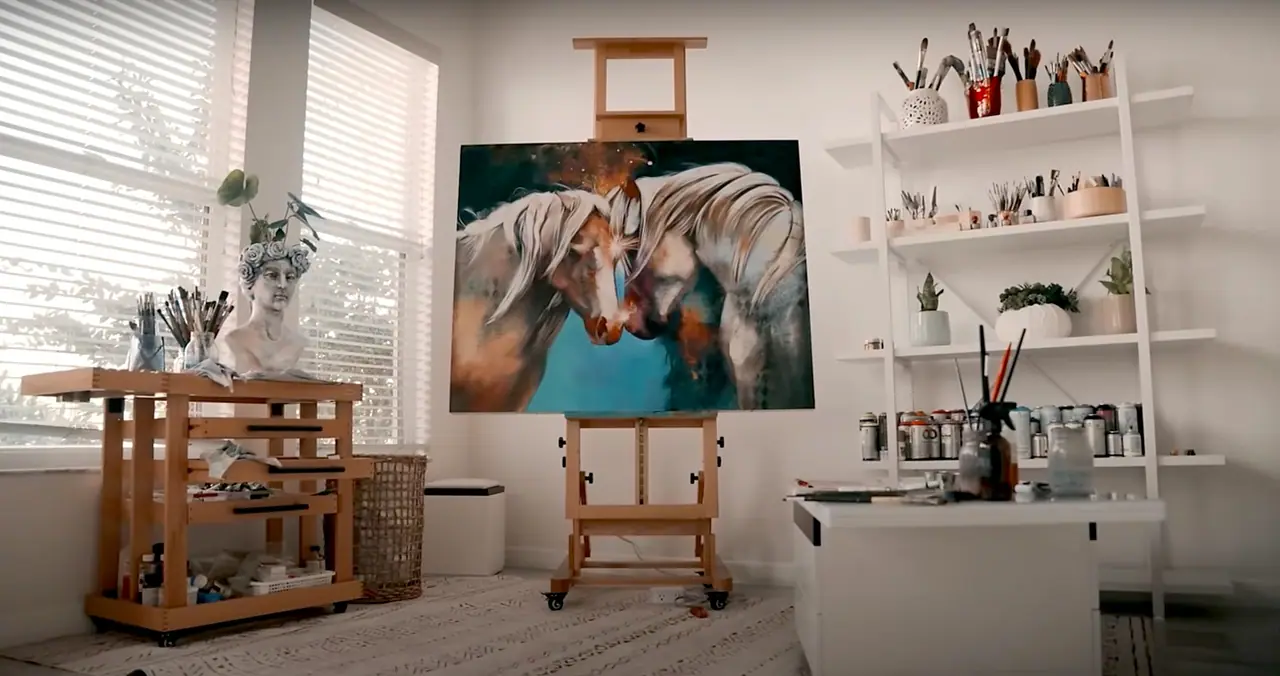 A bright home art studio with an easel, a painting in progress, a work table, and open shelving